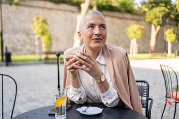 una mujer se toma un café en una terraza. lleva el pelo corto y va vestida con una camisa blanca y una chaqueta de rosa palo de entretiempo