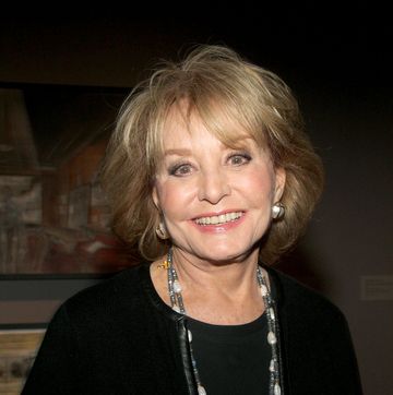 barbara walters smiles at the camera, she is wearing a black outfit with large pearl and gold earrings and a long necklace with multiple strands
