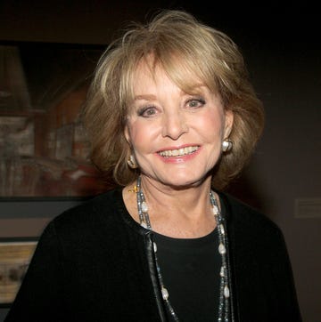 barbara walters smiles at the camera, she is wearing a black outfit with large pearl and gold earrings and a long necklace with multiple strands