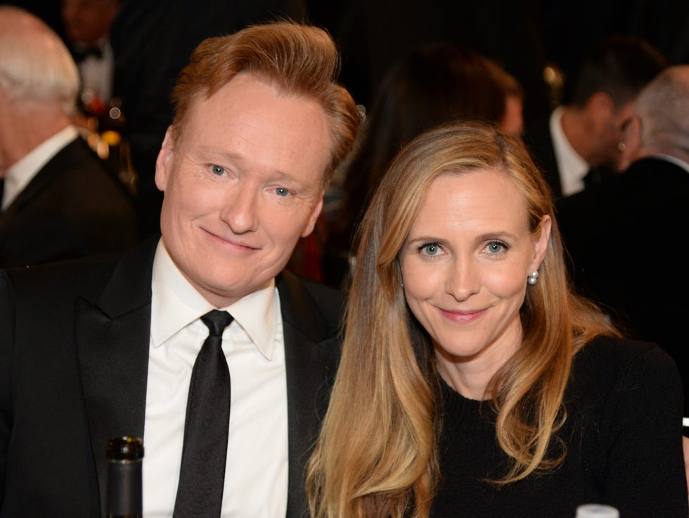 conan o'brien and liza powel o'brien smile at the camera as they sit next to each other, both wear formal black attire