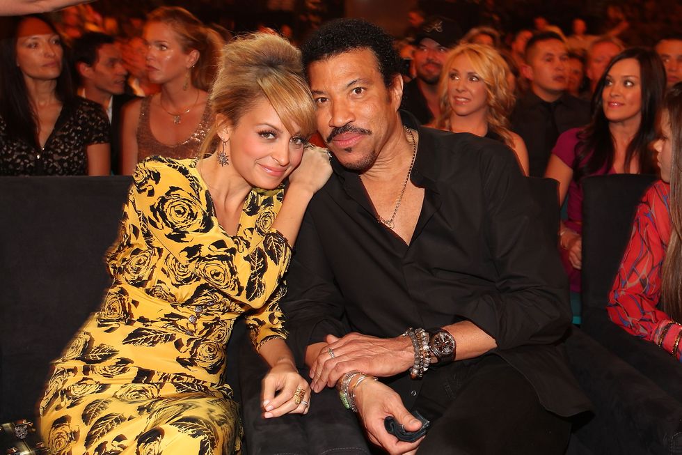 nicole richie and lionel richie lean together while posing for a photo, both look at the camera and smile, nicole wears a yellow and black patterned dress, lionel wears all black with a watch, several bracelets, and a necklace, behind them is a crowd of people