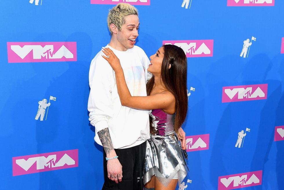 pete davidson, wearing a white shit and black pants, smiles as he receives a hug from ariana grande, who wears a silver dress, as they both stand in front of a blue wall with vma logos on it