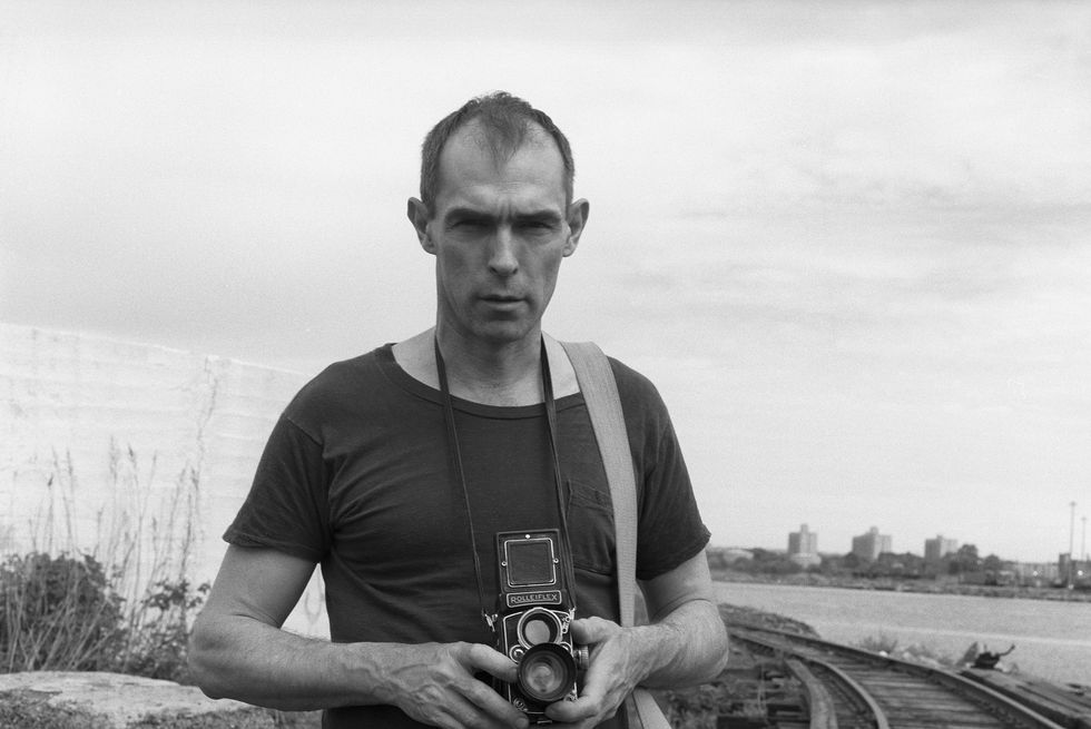 Peter Hujar sostiene una cámara frente a una vía férrea en una imagen en blanco y negro.