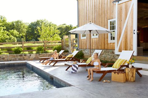 two kids sit on lounge chairs in front of a wooden barn by the pool