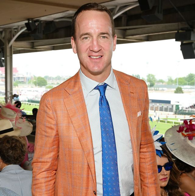peyton manning smiles at the camera, he wears an orange suit jacket, striped collared shirt, and blue tie
