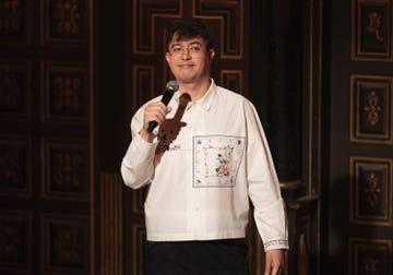 phil wang stands on stage with a microphone for a comedy special with netflix, he has straight black hair and wears glasses, he also wears a white oversized shirt with black trousers