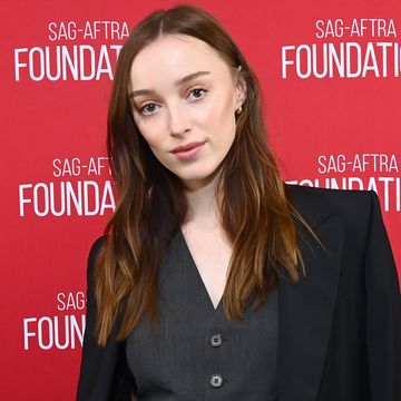 phoebe dynevor, a young woman stands looking at the camera with a neutral facial expression, she has long brown hair worn in loose waves, she wears a dark grey waistcoat and skirt with a black blazer