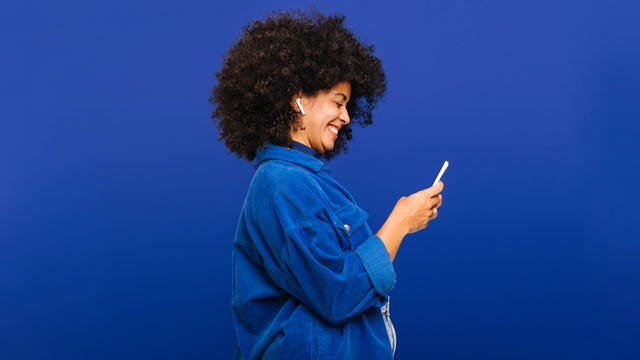 carefree young woman smiling happily while playing music using a smartphone and earbuds. cheerful woman with curly hair enjoying her favourite playlist while standing against a blue background.