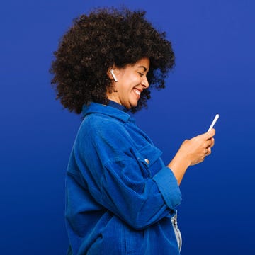 Carefree young woman playing music using a smartphone and earbuds carefree young woman smiling happily while playing music using a smartphone and earbuds. cheerful woman with curly hair enjoying her favourite playlist while standing against a blue background.