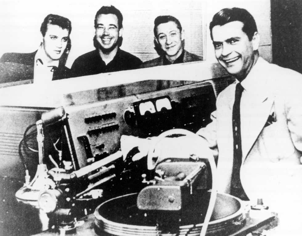 a man smiles while standing at a recording equipment bay, three men stand in the background