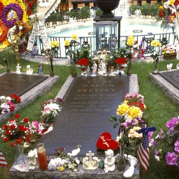 three graves rest in front of a fenced in circular fountain, the graves and entire grounds are decorated with flowers candles and other memorial items