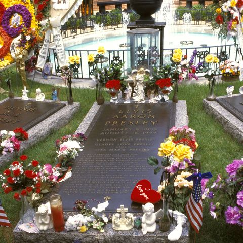 three graves rest in front of a fenced in circular fountain, the graves and entire grounds are decorated with flowers candles and other memorial items