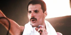 freddie mercury sings into a microphone he holds on a stand and holds one fist out in front of him, he wears red pants, a towel around his neck and a white and red striped wristband