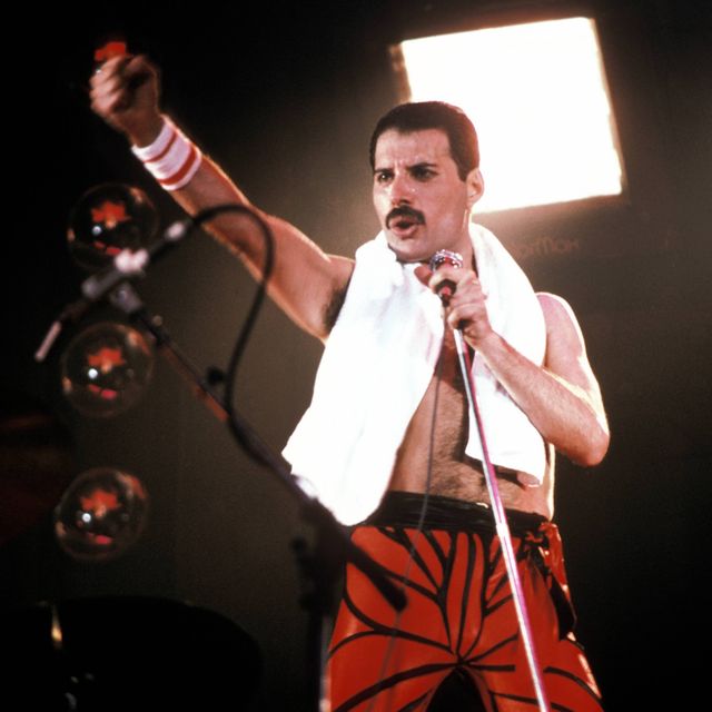 freddie mercury sings into a microphone he holds on a stand and holds one fist out in front of him, he wears red pants, a towel around his neck and a white and red striped wristband