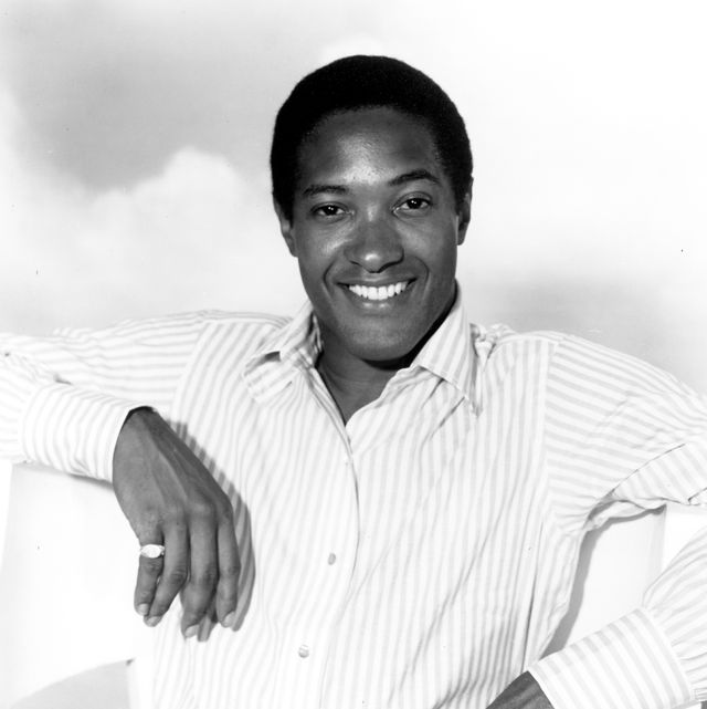 sam cooke smiles at the camera while sitting in a chair with his arms resting on the seat back, he is wearing a striped collared shirt and pinkie ring on his right hand