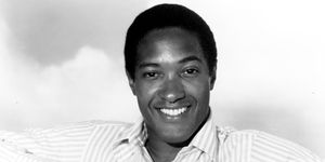 sam cooke smiles at the camera while sitting in a chair with his arms resting on the seat back, he is wearing a striped collared shirt and pinkie ring on his right hand