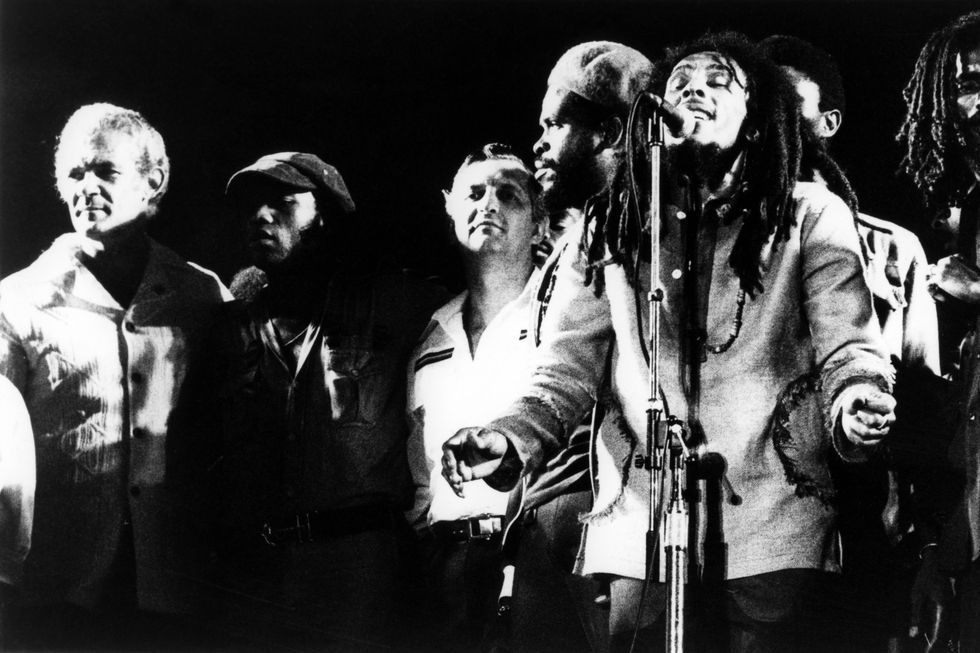 politicians michael manley and edward seaga stand onstage as bob marley sings into a microphone and members of the wailers stand around