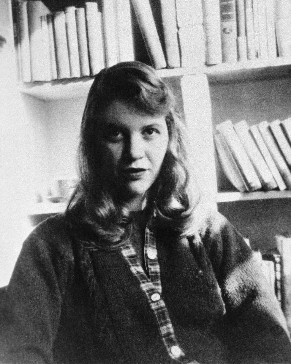 sylvia plath sitting in front of bookshelves and looking ahead for a photograph