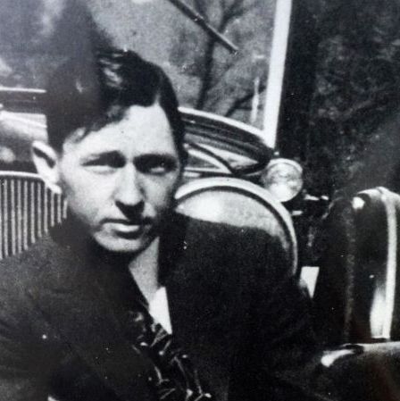 clyde barrow stares at the camera while crouching in front of a car, he holds a gun