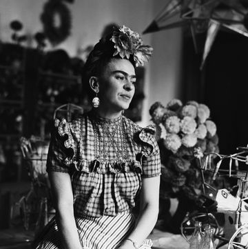 frida kahlo sits on a table while wearing a floral head piece, large earrings, a plaid blouse and striped pants, she looks off to the right