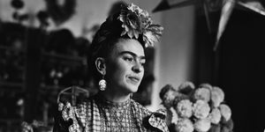 frida kahlo sits on a table while wearing a floral head piece, large earrings, a plaid blouse and striped pants, she looks off to the right