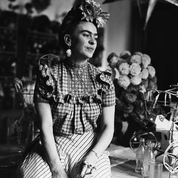 frida kahlo sits on a table while wearing a floral head piece, large earrings, a plaid blouse and striped pants, she looks off to the right