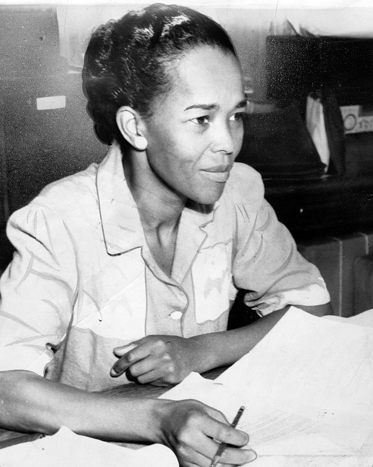 ella baker sits a table with several papers in front of her and holds a pen in one hand, she wears a collared shirt and looks right