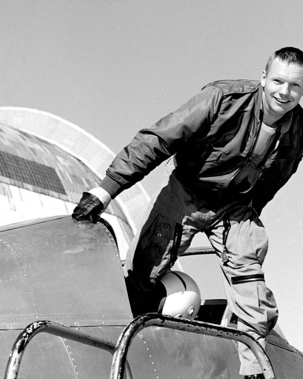 in this black and white photo, a young neil armstrong is standing and stepping out of the cockpit of a plane, he wears a leather jacket, gloves, a light colored shirt, and light colored tactical pants, his right hand touches the frame of the plane just in front of the cockpit as his left foot, likely in a black boot, steps on a platform