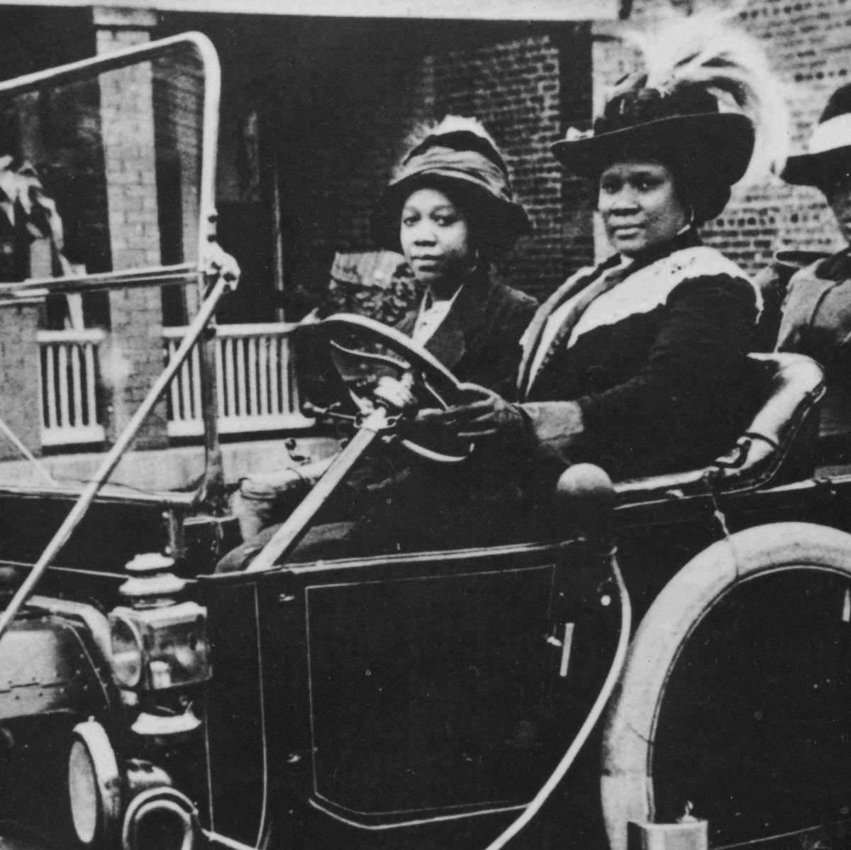 madam cj walker sits in the driver seat of an early car with the top down, a woman sits in the passenger seat, both women wear hats