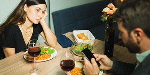 young girlfriend looking annoyed at her busy boyfriend texting on his smartphone during a dinner date