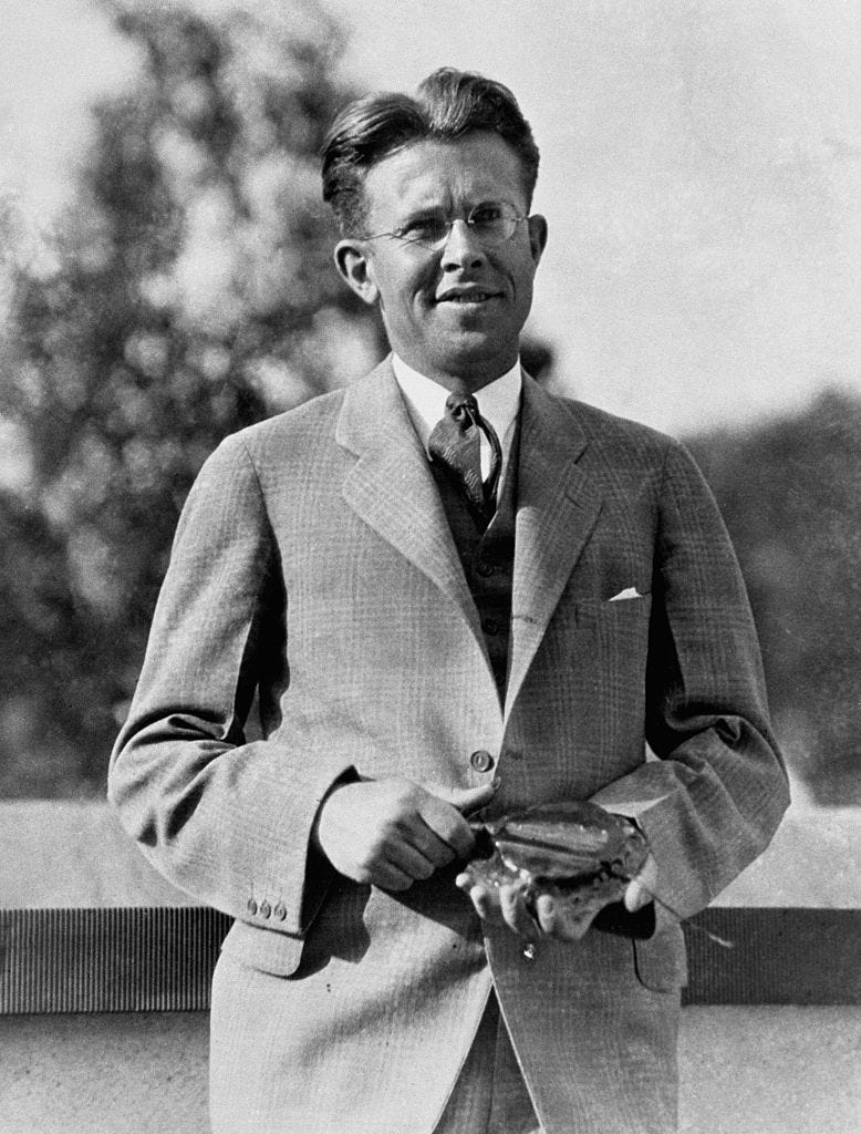 ernest o lawrence stands outside and holds a piece of machinery in his hands, he wears a plaid suit with a light collared shirt and tie