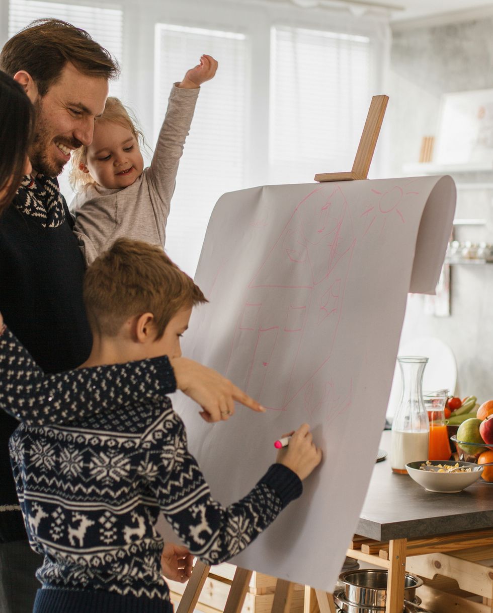 copy space shot of parents bonding and having fun with their teenage son and their toddler daughter they are standing in their living room and drawing on a piece of paper that is hanging on easel