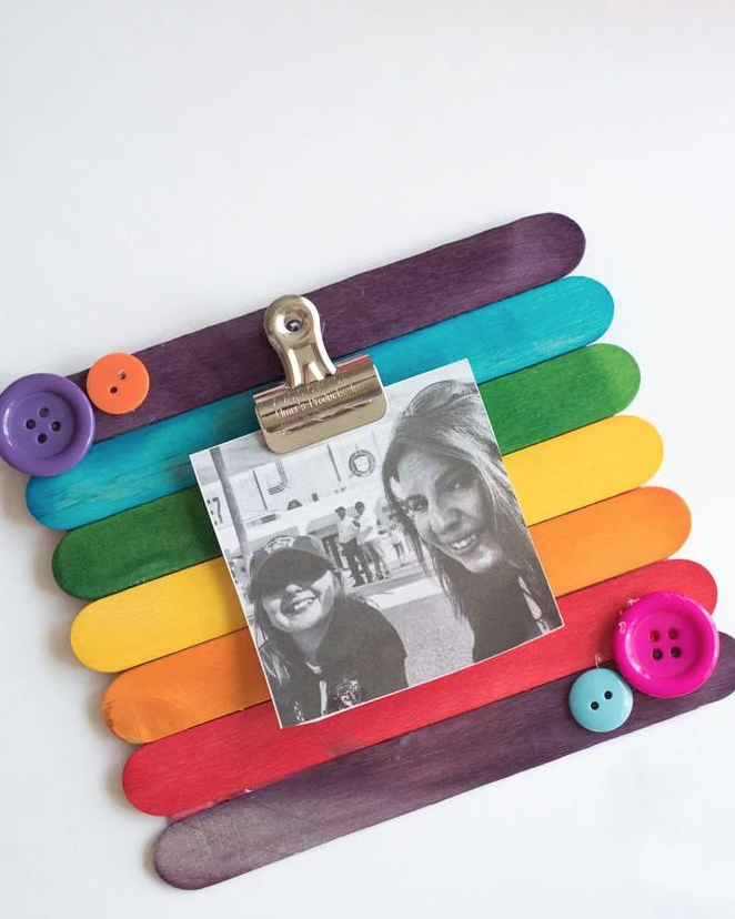 popsicle sticks painted a rainbow of colors glued side by side to form the background of a photo frame, with button decorations and a metal clip attached to hold a small black and white photo of mother and child