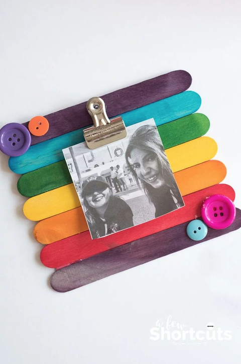 popsicle sticks painted a rainbow of colors glued side by side to form the background of a photo frame, with button decorations and a metal clip attached to hold a small black and white photo of mother and child