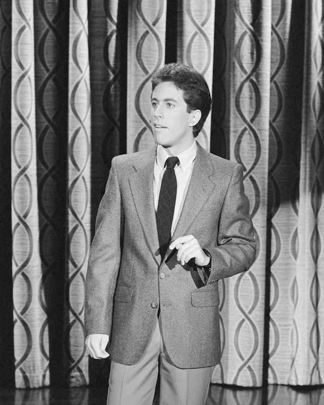 jerry seinfeld stands in front of a patterned curtain in a black and white photo while performing, he wears trousers, a blazer, a collared shirt and tie and looks off camera