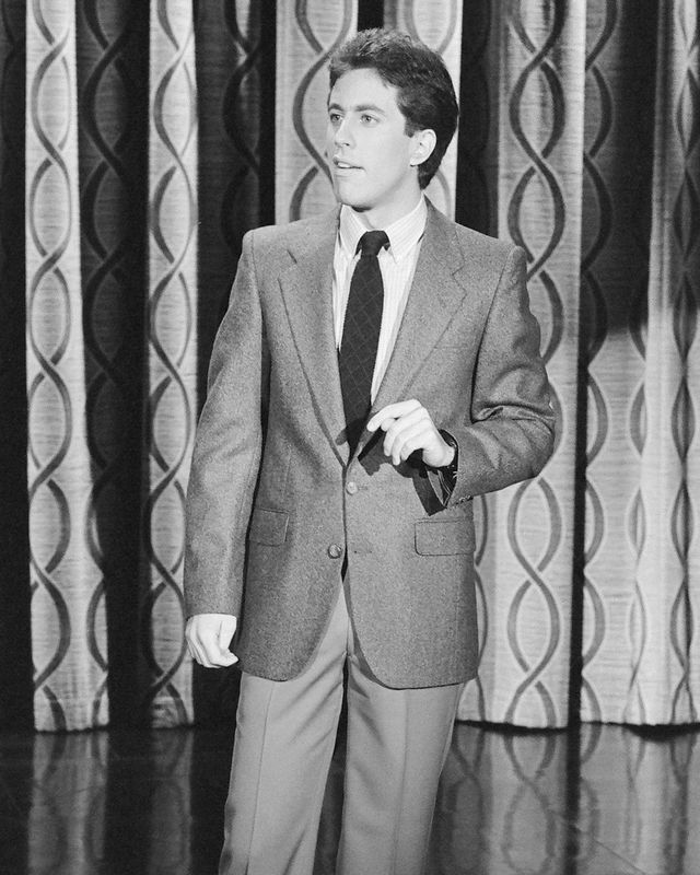 jerry seinfeld stands in front of a patterned curtain in a black and white photo while performing, he wears trousers, a blazer, a collared shirt and tie and looks off camera