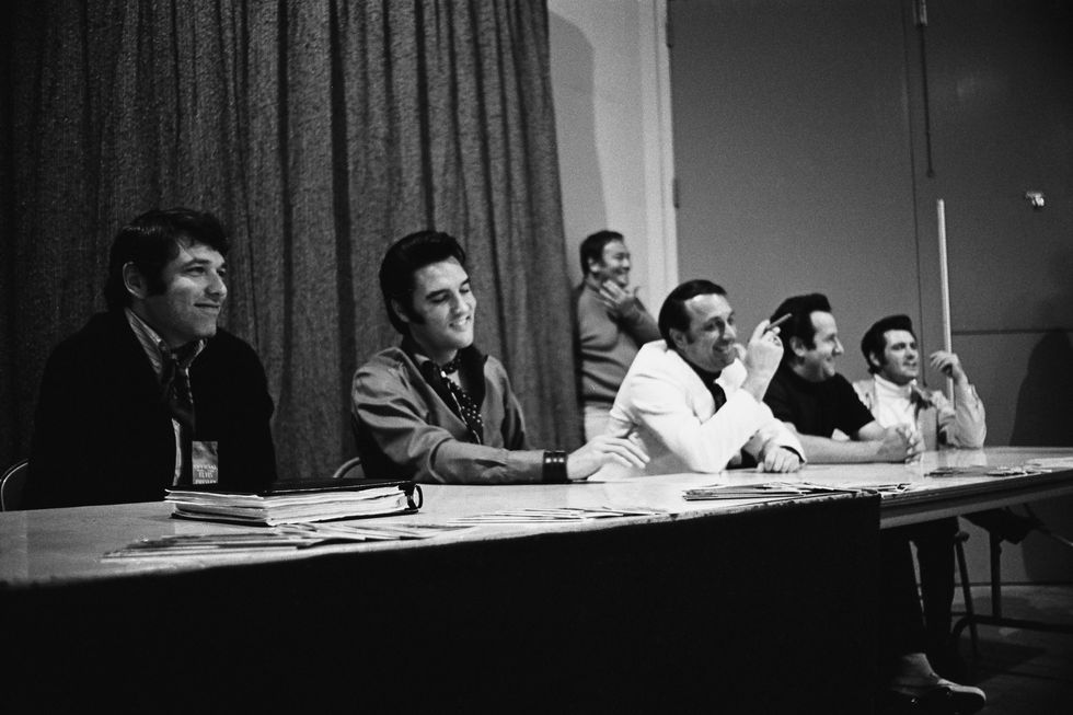 a black and white photo of five men, including elvis presley, sitting at a long table during a press conference and smiling