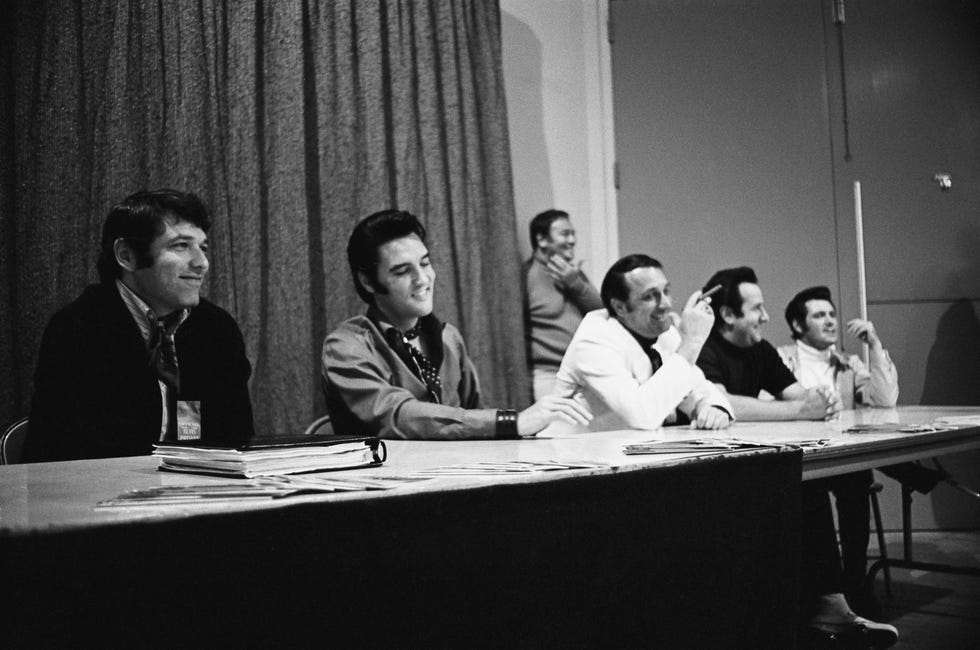 a black and white photo of five men, including elvis presley, sitting at a long table during a press conference and smiling