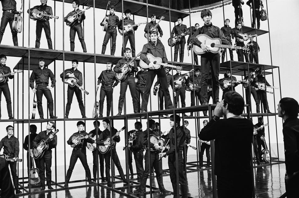a black and white photo of director steve binder yelling up at many extras on a multi level platform, with the extras all holding guitars and dressed like elvis presley
