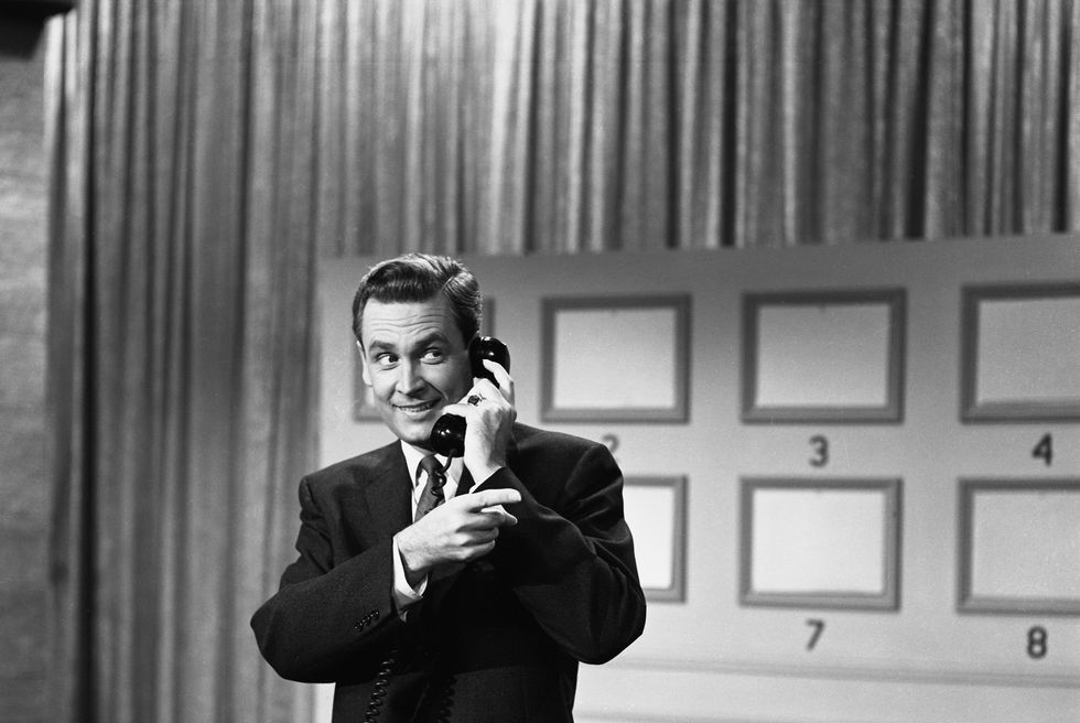 a black and white photo of bob barker on the set of truth or consequences, listening into a telephone and pointing off camera