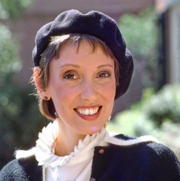 shelley duvall smiles at the camera, she wears a navy beret and matching sweater with a white collared shirt and necklace
