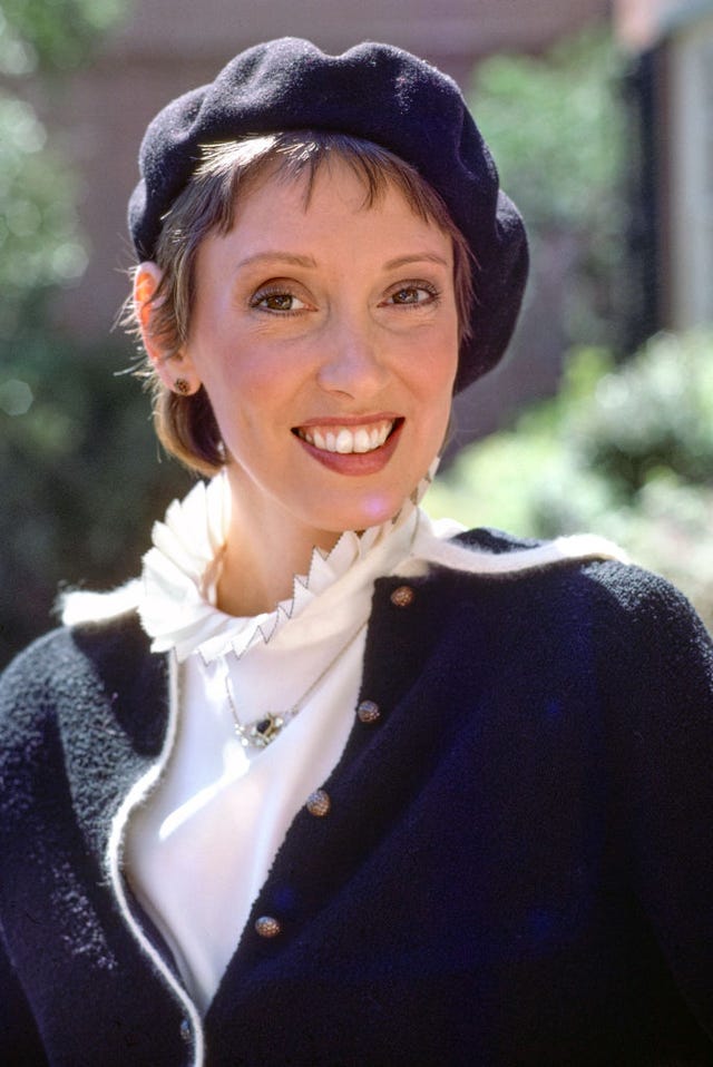 shelley duvall smiles at the camera, she wears a navy beret and matching sweater with a white collared shirt and necklace
