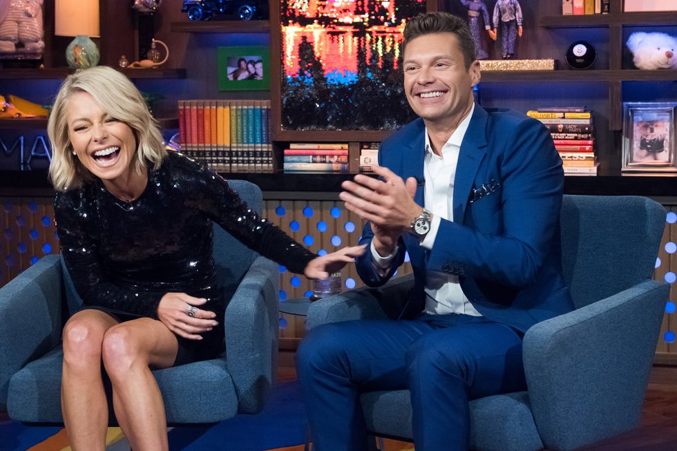 kelly ripa and ryan seacrest sit in armchairs on a tv set, both are laughing and ripa has one arm extended toward seacrest as she leans forward with her eyes closed, seacrest is clapping, ripa wears a sequined black mini dress with long sleeves, seacrest wears a blue suit with a white collared shirt unbuttoned at the neck and a watch