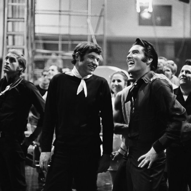 a black and white photo of steve binder and elvis presley laughing and talking behind the scenes of filming while several other people stand behind them