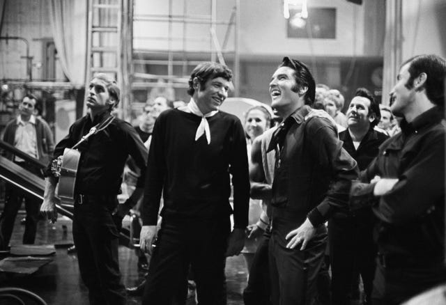 a black and white photo of steve binder and elvis presley laughing and talking behind the scenes of filming while several other people stand behind them