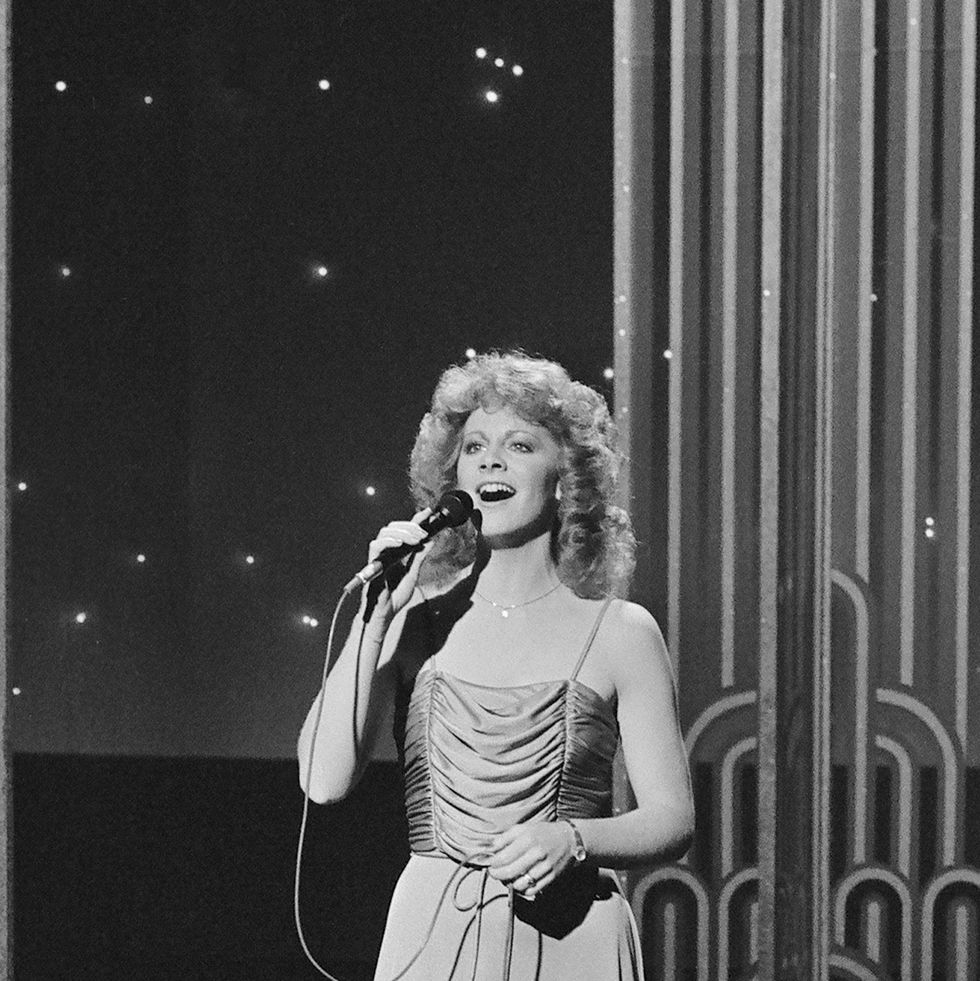 reba mcentire sings into a microphone she holds while standing on a tv set, she wears a dress and a necklace