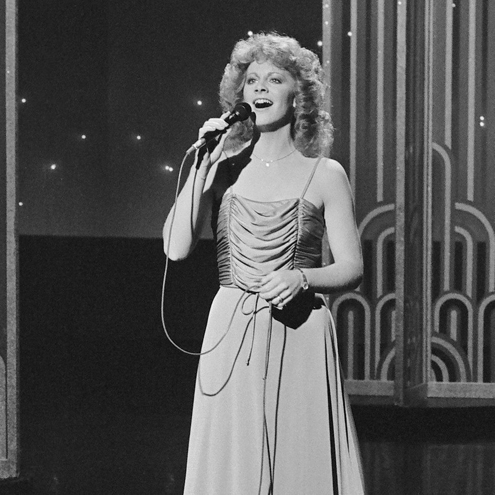 reba mcentire sings into a microphone she holds while standing on a tv set, she wears a dress and a necklace