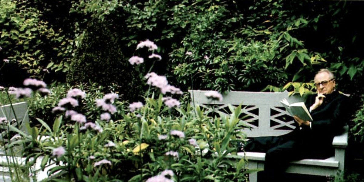 a person sitting on a bench reading a newspaper surrounded by flowers and bushes