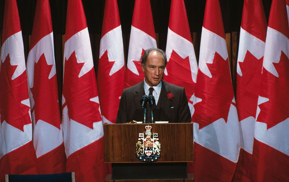 pierre trudeau speaks at a podium, he wears a dark suit with a red rose on his lapel