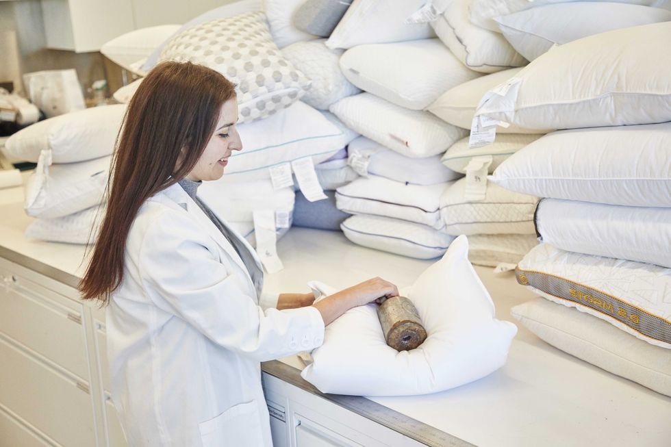 emma seymour, good housekeeping's senior textiles analyst, wearing a white lab coat and adding a weight to a white pillow, surrounded by stacks of white pillows